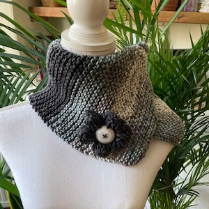 May include: A grey, green and beige knitted cowl with a large grey flower and a button in the centre. The cowl is displayed on a white mannequin.