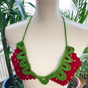 May include: A red and green crocheted bikini top shaped like a strawberry. The top has a green crocheted leaf design around the edges and yellow stitching for the seeds.