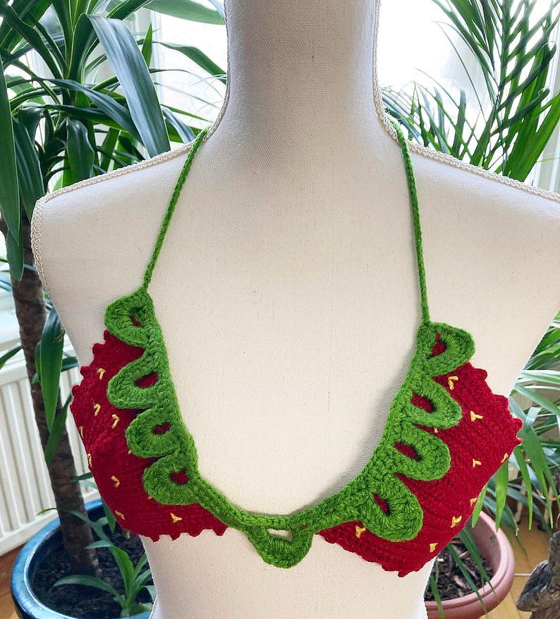 May include: A red and green crocheted bikini top shaped like a strawberry. The top has a green crocheted leaf design around the edges and yellow stitching for the seeds.