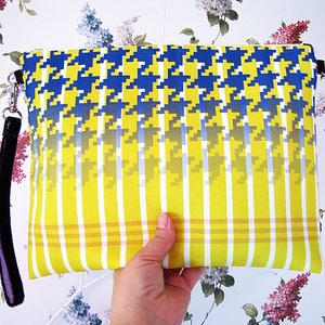 Puede incluir: Clutch rectangular con un estampado de pata de gallo en azul y amarillo, que transiciona a rayas verticales amarillas y blancas. Tiene una correa de muñeca negra y herrajes plateados. La bolsa se sostiene contra un fondo de papel pintado floral.