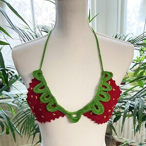 May include: A red and green crocheted bikini top shaped like a strawberry. The top has a green crocheted leaf design and a red crocheted strawberry design.