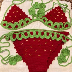 May include: A red and green crocheted bikini set shaped like a strawberry. The bikini top has a halter style with a green crocheted flower detail. The bikini bottom has a high-waisted style with a green crocheted trim.