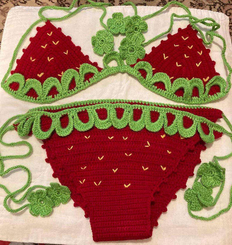 May include: A red and green crocheted bikini set shaped like a strawberry. The bikini top has a halter style with a green crocheted flower detail. The bikini bottom has a high-waisted style with a green crocheted trim.
