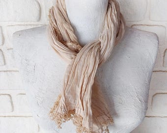 May include: A beige scarf with a delicate lace trim is draped around a white mannequin. The scarf has a crinkled texture and is displayed against a white brick wall. The scarf is a neutral colour.