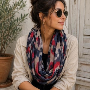 Peut inclure: Une écharpe infinie arborant un motif ikat répétitif dans des tons bleu marine, rouge profond et beige. L'écharpe est portée sur un haut noir et une veste beige clair, accompagnée de créoles dorées et de lunettes de soleil rondes foncées.
