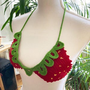 May include: A red and green crocheted bikini top shaped like a strawberry. The top has a green crocheted leaf design around the edge.