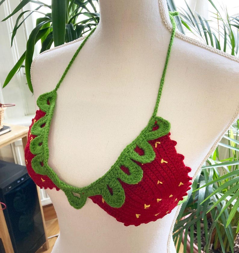 May include: A red and green crocheted bikini top shaped like a strawberry. The top has a green crocheted leaf design around the edge.