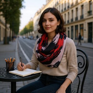 May include: A woman wearing a beige sweater and dark jeans, seated at a black table, writing in a notebook. She wears a scarf with red, black, and pink abstract designs. A pencil holder with pencils sits on the table.