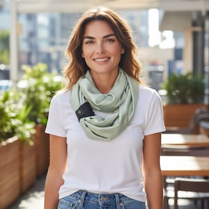 May include: A light green infinity scarf with a black clasp, worn by a person. The scarf is wrapped around the neck. The person is wearing a white t-shirt and blue jeans. The background is blurred, suggesting an outdoor location.