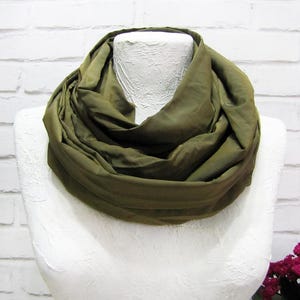 Puede incluir: Bufanda infinita verde oliva drapeada sobre un maniquí blanco. La bufanda está hecha de una tela suave y fluida y está envuelta varias veces alrededor del cuello. El fondo es una pared de ladrillos blancos.