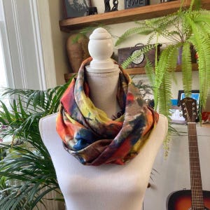 Puede incluir: Bufanda infinita multicolor exhibida en un maniquí blanco. La bufanda presenta un diseño abstracto vibrante con tonos de rojo, naranja, amarillo y azul. La tela es suave y fluida.