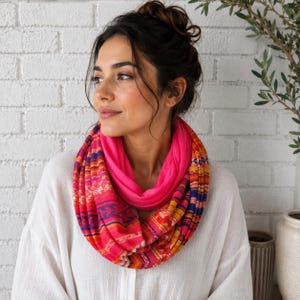 Peut inclure: Une écharpe infinie présentant un motif vibrant de rayures horizontales dans des tons de rose vif, orange, jaune et bleu profond. L'écharpe est portée autour du cou, révélant une couche intérieure rose vif unie et un tissu extérieur multicolore.