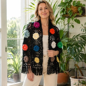 May include: A black crochet cardigan featuring a grid pattern and scattered three-dimensional floral motifs in various colors including red, yellow, blue, and green. The garment has long sleeves and a hem finished with long black fringe tassels.