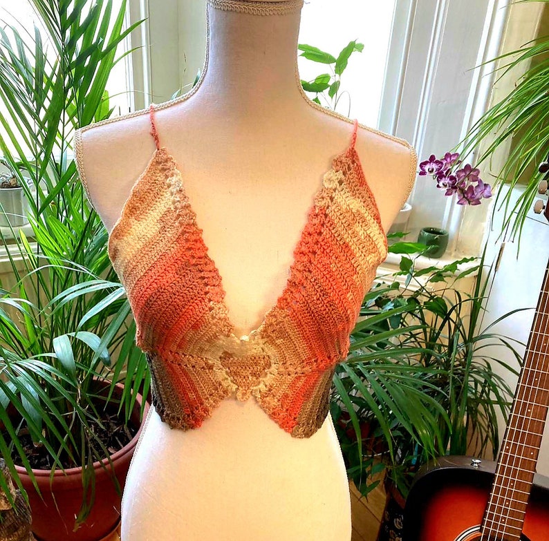 May include: A crocheted halter top with a pink, orange, and beige ombre pattern. The top has a V-neckline and a scalloped edge.