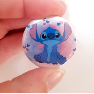 Peut inclure: Un badge rond de 32 mm représentant le personnage Stitch du film Lilo & Stitch. Le personnage bleu est placé sur un fond aquarelle rose et bleu avec des cœurs. Le texte "Badge 32mm STITCH" est en bas.