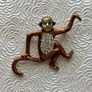 May include: A bronze-colored monkey brooch with a gold-colored head and green eyes. The monkey's chest is covered in clear rhinestones. The brooch is set against a white, textured background.
