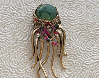 May include: A gold-toned jellyfish brooch featuring a large, smooth, dark green cabochon stone as the head. The tentacles are adorned with small, sparkling red, black, and clear rhinestones set into the textured metal surface of the creature.