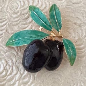 May include: A gold-toned brooch featuring two glossy black olives and three teal green leaves. The leaves have white veins and are attached to the olives with a gold stem. The brooch is set against a textured, off-white background.