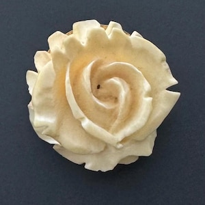 May include: A cream-coloured rose brooch with detailed petals. The brooch is made of a light-coloured material and has a gold-coloured metal backing. The rose is the focal point of the image.