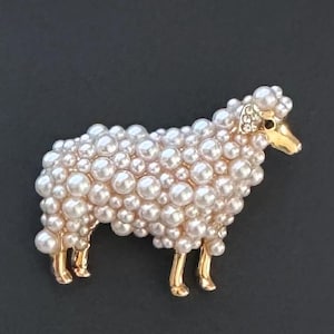 Puede incluir: Broche con forma de oveja con un marco de metal dorado. El cuerpo de la oveja está cubierto de perlas de imitación, y la cabeza presenta un pequeño grupo de cristales transparentes. El broche tiene patas doradas y un hocico dorado.