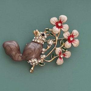 May include: A detailed brooch featuring a squirrel holding a branch of flowers. The squirrel is brown and gray with a jeweled crown. The flowers are white with red centers and green leaves. The brooch is gold-toned.