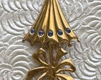 May include: Gold-toned umbrella brooch with a bow detail. The umbrella top features six blue gemstones. The brooch is a vintage accessory, perfect for adding a touch of elegance to any outfit. The brooch is made of metal.