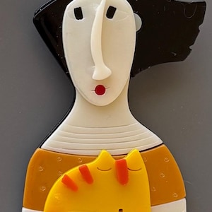 May include: A whimsical brooch featuring a stylized woman with a black hairstyle, a white face, and a red lip. The woman is wearing an orange top and is embracing a yellow cat with orange stripes.