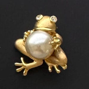 Op de afbeelding: Een goudkleurige kikkerbroche met een grote, ronde parel in zijn armen. De kikker heeft twee kleine, heldere edelstenen als ogen. De broche staat tegen een zwarte achtergrond.