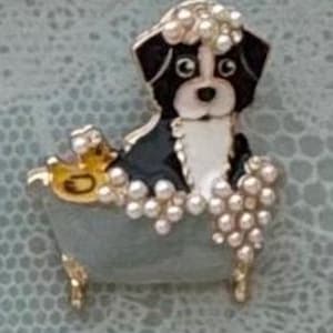 May include: A gold-tone metal brooch featuring a black and white dog sitting in a light blue clawfoot bathtub. The dog is covered in white faux pearl bubbles, with additional pearl bubbles resting on the edge of the tub and a small yellow duck.