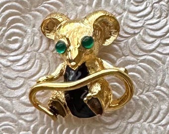 May include: Gold-toned koala bear brooch with green gemstone eyes and a black enamel belly. The koala is holding its tail, creating a circular design. The brooch is set against a textured, off-white background.