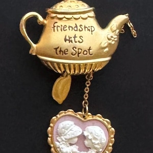May include: A gold-colored teapot-shaped brooch with the words "friendship hits the spot." A heart-shaped pendant with two white cameo figures hangs below. The pendant has a pink background and a gold frame. The brooch is on a black background.