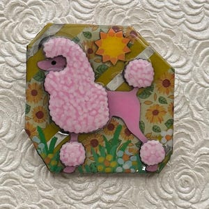 May include: A pink poodle design on a decorative, multi-sided piece. The poodle has a textured coat and is set against a background of sunflowers and a yellow sun. The piece is surrounded by a green and yellow border.