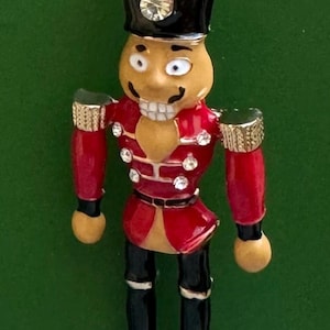 May include: A nutcracker brooch featuring a red jacket with clear rhinestones, black boots, and a black hat adorned with a gold-tone plume and a single clear rhinestone. The figure has a tan face with white teeth and black painted eyes.