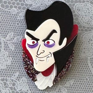 May include: A vampire brooch featuring a pale face with sharp fangs, purple eyeshadow, and red eyes. The character has black hair, a high-collared black cape with red lining, and a white cravat, set against a dark, glittery, textured background.