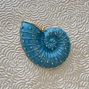Könnte beinhalten: Eine Nautilus-Muschel-Brosche mit glänzender blauer Emaille-Oberfläche und marmorierter Textur. Die Muschel ist mit zahlreichen kleinen, klaren, runden Strasssteinen verziert, die entlang der spiralförmigen Rippen und am Goldrand sitzen.