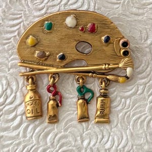 May include: A gold-toned artist's palette brooch with a variety of paint colors. Two crossed paintbrushes sit atop the palette. Dangling below are miniature bottles and tubes labeled "Linseed Oil" and "White".