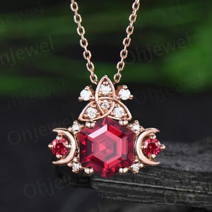 May include: A rose gold necklace with a pendant featuring a large, faceted, red gemstone. The pendant is adorned with smaller white gemstones and crescent moon accents, creating an elegant and intricate design.