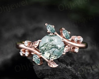 May include: A rose gold ring with a green moss agate gemstone surrounded by delicate leaf-shaped accents and small clear gemstones.