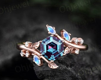 Puede incluir: Un anillo de oro rosa con una piedra preciosa de alexandrita hexagonal rodeada de pequeñas piedras blancas y acentos de piedra lunar. El anillo presenta un diseño delicado y de hojas.
