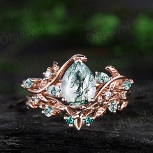 May include: A rose gold engagement ring with a pear-shaped green moss agate gemstone surrounded by smaller green and white gemstones. The ring features a delicate leafy design.