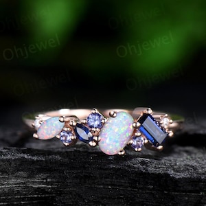 May include: A delicate rose gold band ring with a cluster of white opal and blue sapphire gemstones. The ring features a variety of shapes and sizes of gemstones, creating a unique and eye-catching design.