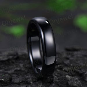 May include: A close-up shot of a sleek, black ring. The ring has a smooth, polished surface that reflects light. The ring is set against a dark, textured background, highlighting its glossy finish. The ring appears to be made of a solid, dark material.