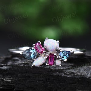 May include: A silver ring with a white opal, pink and blue gemstones. The ring is set on a dark gray background.