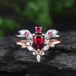 May include: A silver and rose gold ring set featuring a pear-shaped ruby gemstone surrounded by smaller diamonds. The top ring has a bat design with wings and the bottom ring has a crescent moon and star design.