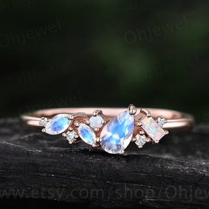 May include: A delicate rose gold ring with a cluster of moonstone and opal gemstones. The ring features a crescent shape with a variety of sizes and shapes of gemstones.