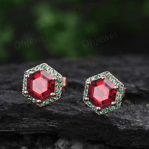 May include: A pair of red gemstone earrings with a hexagonal shape. The earrings are set in rose gold and have a halo of smaller green gemstones.