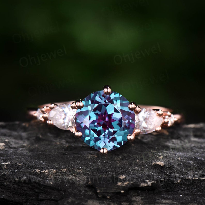 Pode incluir: Um anel de noivado em ouro rosa com uma grande pedra de alexandrita redonda azul e roxa cercada por dois diamantes menores em forma de p&ecirc;ra.