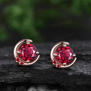 May include: A pair of crescent moon earrings with a red gemstone in the center and smaller red gemstones on the sides. The earrings are made of rose gold.