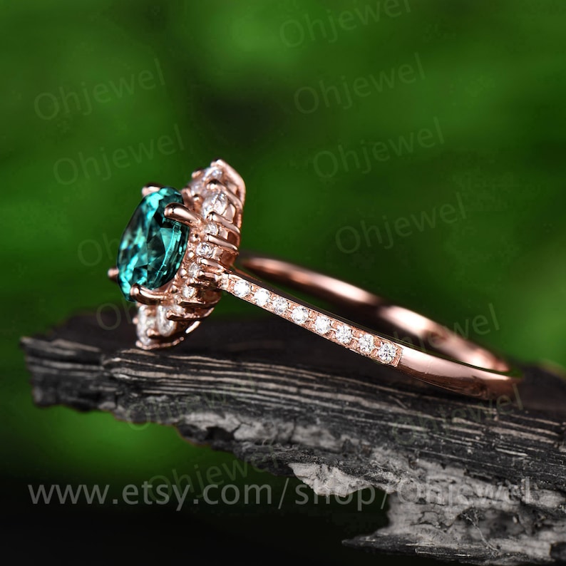 May include: A rose gold engagement ring with a large oval green gemstone surrounded by smaller round white gemstones. The ring has a band that is also set with smaller round white gemstones.