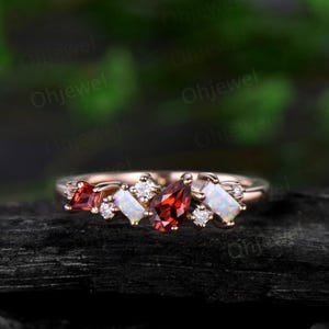May include: A rose gold ring with a pear-shaped garnet, two rectangular opals, and smaller round diamonds. The ring is set on a black background.